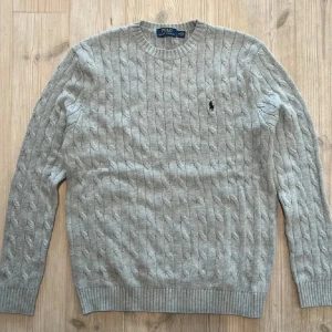 Grå kabelstickad tröja från Polo Ralph Lauren - Säljer en klassisk grå kabelstickad tröja från Polo Ralph Lauren med rund halsringning och broderad logga på bröstet. Perfekt för en stilren och avslappnad look. Pris kan diskuteras vid snabb affär