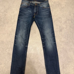 Mörkblå jeans från Nudie Jeans - Säljer ett par mörkblå jeans från Nudie Jeans med klassisk femficksdesign och orangea sömmar. Jeansen har normal passform och raka ben, perfekta till vardags. 28/32