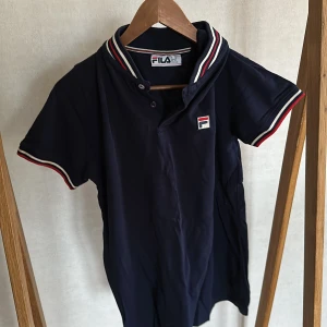 Mörkblå pikétröja från FILA - Säljer en klassisk mörkblå pikétröja från FILA med korta ärmar. Tröjan har vit, röd och marinblå rand på krage och ärmslut samt FILA-logga på bröstet.