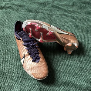 Nike Mercurial fotbollsskor i kopparfärg - Säljer ett par Nike Mercurial fotbollsskor i kopparfärg med svart snörning och vit Nike-logga. Skorna har hög krage och dobbar för spel på gräs. Perfekta för dig som vill ha stilrena och snabba skor på planen.