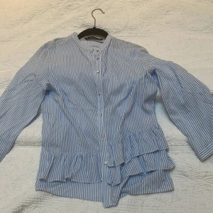 Blårandig blus med volang från Zara Basic - Säljer en blårandig blus från Zara Basic i storlek XS. Blusen har långa ärmar, knappar framtill och volangdetaljer nedtill. Tillverkad i mjuk bomull och har en klassisk, avslappnad passform. Perfekt för dig som gillar stilrena ränder och fina detaljer.