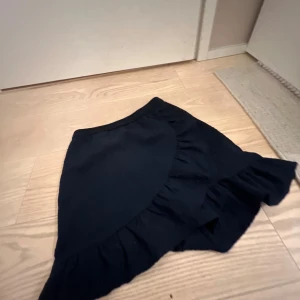 Svart volangkjol från ONLY - Säljer en svart kortkjol från ONLY med volangdetalj längs kanten. Kjolen har en ribbad struktur och resår i midjan för en bekväm passform. Perfekt för dig som gillar en enkel men ändå trendig stil.