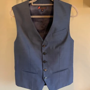 Mörkblå kostymväst med knappar - Säljer en stilren mörkblå kostymväst från Dressmann i storlek XS. Västen har klassisk passform, tre fickor framtill och knäppning med fem knappar. Justerbart band i ryggen för perfekt passform. Perfekt till kostym eller som en snygg detalj till skjorta.