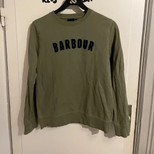 Olivgrön sweatshirt från Barbour - Säljer en olivgrön sweatshirt från Barbour med svart text på bröstet. Tröjan har rund halsringning och långa ärmar. Perfekt för en avslappnad stil och enkel att matcha med jeans eller joggers. 