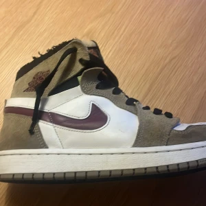 Nike Air Jordans i vitt och brunt - Säljer ett parAir Jordan 1 Zoom CMFT "Neutral Olive" med vit läderbas, bruna mockadetaljer och svarta skosnören. Skorna har en mörkröd swoosh och högt skaft. Perfekta för dig som gillar streetwear och vill sticka ut med klassiska sneakers. Säljer skorna pågrund av att den är för små. Pris kan diskuteras.