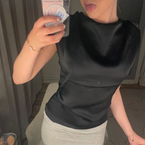 Svart satinblus - Snygg svart blus i satin med markerade axlar och ärmlös design. Blusen har en elegant, slät yta och rund halsringning. Perfekt för att lyfta vilken outfit som helst med en stilren touch.