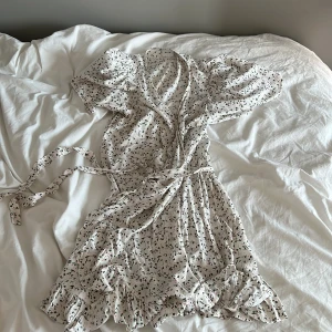 Vit blommig omlottklänning från Vila - Supersöt vit omlottklänning från Vila med små blommor i grönt och rosa. Klänningen har korta puffärmar, volangkant nertill och knyts i midjan. Perfekt för sommarens alla tillfällen!