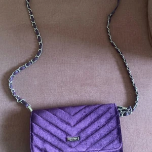 Lila axelväska i sammet från River Island - Säljer en lila axelväska i sammet från River Island med kedjerem och quiltad design. Väskan har ett fack med dragkedja och flera kortfack på insidan. Perfekt för dig som vill ha en snygg accessoar med plats för det viktigaste.