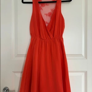 Röd omlottklänning med spetsdetaljer - Snygg röd klänning med omlott framtill och djup v-ringning. Baksidan har transparent spets med blommigt mönster. Klänningen har ärmlös design och markerad midja, perfekt för sommarens tillfällen.