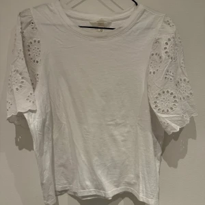 Vit topp med broderade puffärmar - Säljer en vit topp med korta puffärmar i broderad bomull med hålmönster. Toppen har rund halsringning och är i mjukt material. Perfekt för en fräsch och söt look!