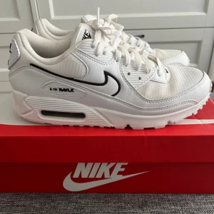 Nike Air Max 90 vita sneakers - Säljer ett par fräscha vita Nike Air Max 90 sneakers med svart swoosh. Skorna har klassisk design med snörning, synlig Air Max-dämpning och platt sula. Perfekta för dig som gillar stilrena och sportiga skor.