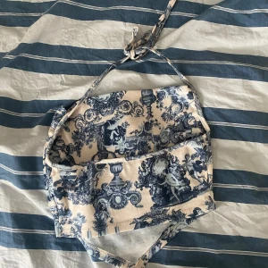 Blåvit topp med vintage-mönster - Säljer en unik blåvit topp med klassiskt vintage-inspirerat mönster. Toppen har ribbad struktur och knytband både upptill och i midjan. Toppen har en v-ringad form. Aldrig använd 