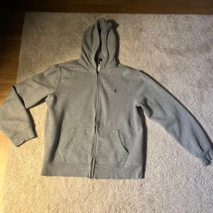 Grå hoodie från Polo Ralph Lauren - Säljer en klassisk grå hoodie från Polo Ralph Lauren i storlek XL barnstorlek. XL i barnstorlek motsvara i vuxenstorlek XS/S. Tröjan har dragkedja, huva och två fickor framtill samt den ikoniska broderade loggan på bröstet. Perfekt för en avslappnad stil.
