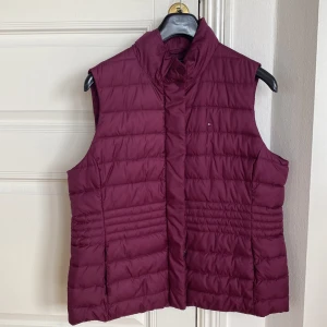 Vinröd dunväst från Tommy Hilfiger - Säljer en vinröd dunväst från Tommy Hilfiger. Västen har hög krage, quiltad design och stängs med dolda tryckknappar. Perfekt för lager-på-lager och ger en sportig look. Mörkare och snyggare i verkligheten. Sparsamt använd. Stl XL. 