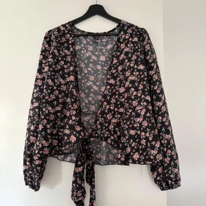 Svart blommig omlottblus  - Säljer en svart omlottblus från Dazign med rosa och vita blommor. Blusen har långa ärmar med resår vid ärmslut och knytband framtill. Endast testad så aldrig använd 