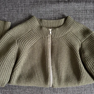 Olivgrön stickad cardigan jacka med dragkedja - Säljer en olivgrön stickad cardigan jacka med grov ribbad struktur och silverfärgad dragkedja framtill. Jackan har lång ärm och en hög, rundad krage. Perfekt för lager-på-lager och enkel att matcha med olika outfits.