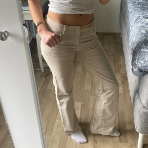 Beiga manchesterbyxor med bootcut - Snygga beiga manchesterbyxor med bootcut och hög midja. Byxorna har klassiska fickor fram och bak samt stängs med dragkedja och knapp. Perfekta för dig som gillar retrostil och vill ha ett par byxor som sticker ut lite extra.
