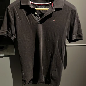 Svart pikétröja från Tommy Hilfiger str 170 cm - Klassisk svart pikétröja från Tommy Hilfiger med liten broderad logga på bröstet och krage med knappar. Perfekt för en stilren och avslappnad look. Tillverkad i bomull för skön komfort.