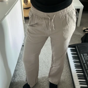 Beige linnebyxor från 157 - Säljer ett par beige linnebyxor från 157 med elastisk midja och dragsko. Byxorna har raka ben och ficka bak, perfekta för en avslappnad stil. Lätta och luftiga, passar bra till varmare dagar.