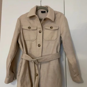 Beige kappa med bälte och knappar - Säljer en beige overshirt med klassisk krage, knappar framtill och två bröstfickor. Jackan har ett matchande bälte i midjan för en snygg siluett. Perfekt för lager-på-lager och enkel att styla.