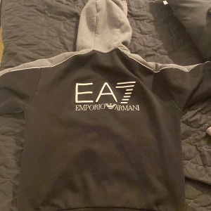 Svart hoodiejacka från Emporio Armani EA7 - Svart hoodiejacka från Emporio Armani EA7 med grå huva och axelparti. Jackan har dragkedja framtill, två fickor med dragkedja och stor logga på ryggen. 