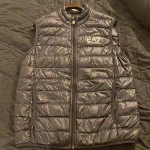 Svart dunväst från EA7 Emporio Armani - Säljer en svart, quiltad dunväst från EA7 Emporio Armani med dragkedja framtill och logga på bröstet. Västen har hög krage och är lätt vadderad, perfekt för lager-på-lager-stil.