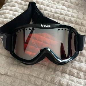 Svarta skidglasögon från Bollé - Snygga svarta skidglasögon från Bollé med bred elastisk rem och spegellins i rödton. Perfekta för vinterns äventyr i backen. Klassisk design med ventilerad ram och bekväm passform. Aldrig använda 