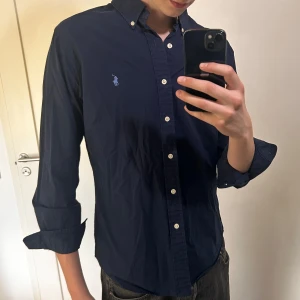 Mörkblå skjorta från Ralph Lauren - Säljer en klassisk mörkblå skjorta från Ralph Lauren med button-down krage och broderad logga på bröstet. Skjortan har långa ärmar och vita knappar framtill. Perfekt för en stilren look. Storlek L slimfit men funkar för M