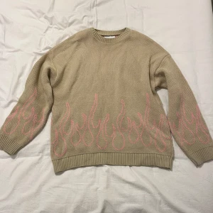Beige stickad tröja med rosa flames från ASOS DESIGN - Om att bli kallad Rosa Flamman av ditt kompisgäng står på din bucket list 2025 så har du hitta helt rätt. Perfekt för att visa din dominans gentemot dina skolkamerater, familj eller opposition, fungerar i alla lägen. 