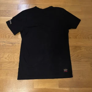 Svart t-shirt från Paul & Shark - Svart t-shirt från Paul & Shark med liten haj-logga på bröstet och tryckt text på ärmen. Klassisk rund hals och normal passform. Perfekt för en clean och stilren look.