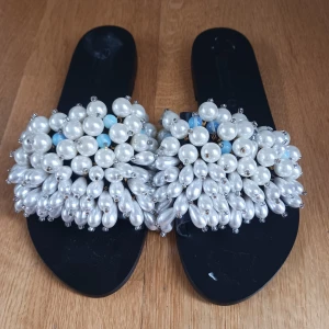 Elina Linardaki tofflor - Unika svarta sandaler med bred rem täckt av stora vita pärlor och några ljusblå pärlor. Platt sula och öppen tå. Perfekta för att sticka ut med en lyxig och trendig look. Storlek 40. Helt oanvända. Märkena på sulan som syns på bilden är endast klister från prislappen som går att då bort enkelt. Nypris 2500kr då dom är handgjorda i Grekland. Pris går att diskutera.