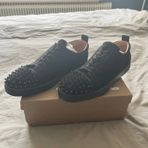 Svarta sneakers med nitar från Christian Louboutin - Säljer ett par svarta sneakers från Christian Louboutin med svarta nitar på tån och längs sidorna. Skorna har snörning och är tillverkade i mocka med läderdetaljer invändigt. Kommer med originalkartong, dustbag och äkthetsbevis.