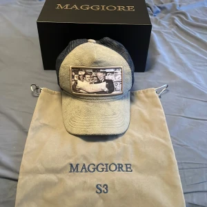 Beige och svart keps från Maggiore - Snygg keps från Maggiore i beige framtill och svart mesh baktill. Framsidan har ett stort broderat motiv med vintage-foto. Justerbar passform med klassisk kepsform. Kommer i exklusiv förpackning och dustbag.