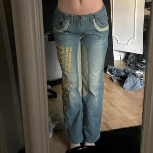 Unika blå jeans med tryck och kontrastsömmar - Säljer ett par blå jeans med raka ben, coola kontrastsömmar och gult tryck längs ena benet. Jeansen har klassiska fickor fram och bak samt en bredare linning i ljusare denim. Perfekta för dig som vill sticka ut med din stil!