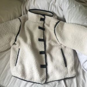 Vit teddyjacka med svarta detaljer - Mysig vit teddyjacka med svarta kantband och stängning framtill. Jackan har hög krage och långa ärmar, perfekt för kyliga dagar. Två fickor framtill och en unik design med svarta detaljer längs kanterna.
