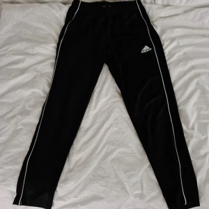 Svarta träningsbyxor från Adidas med vita ränder stl L - Svarta träningsbyxor från Adidas med vita ränder längs benen och broderad logga på låret. Byxorna har en sportig look och är perfekta för träning eller chill. Tillverkade i syntetmaterial för extra komfort.