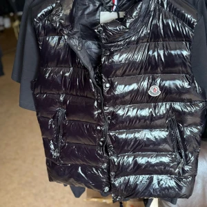 Svart dunväst från Moncler - Säljer en glansig svart dunväst från Moncler med klassisk logotyp på bröstet och dragkedja framtill. Västen har två fickor med dragkedja och ett unikt serietryck på insidan. Perfekt för lager-på-lager-stil. Finns även i alla storlekar hör av er