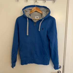 Blå zip hoodie från U.S. Polo Assn. - Säljer en blå hoodie från U.S. Polo Assn. med dragkedja och grå insida på huvan. Tröjan har två fickor framtill, justerbara snören i huvan och broderad logga på bröstet. Perfekt för en avslappnad stil. Inga som helst defekter hör av sig vid minsta fråga!