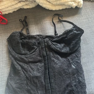 Svart spetsig korsettopp - Säljer en svart korsettopp med tunna justerbara axelband och vacker spets över hela toppen. Toppen har markerade kupor och en dekorativ söm framtill. Perfekt för att lyfta en outfit med en elegant touch. Stolek 75B