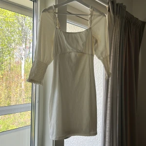 Vit kortklänning med puffärmar och knytning - Säljer en söt vit kortklänning med puffiga trekvartsärmar och smock vid ärmsluten. Klänningen har en fin knytning baktill och lätt rynkade detaljer vid axlarna. Perfekt för varma dagar och somriga tillfällen. OBS finns fläckar, ej försökt få bort dem.