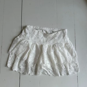 Vit spetskjol med volang - Supersöt vit kjol i spets med volang och bred midja. Kjolen har ett romantiskt uttryck med blommigt spetsmönster och är perfekt för dig som gillar en feminin stil. Använt 1 gång.