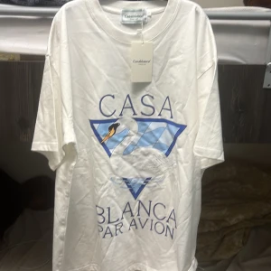 Vit t-shirt med svan-tryck från Casablanca - Vit t-shirt från Casablanca Tennis Club med stort tryck framtill: en svan och blå trianglar samt texten 'Casa Blanca Par Avion'. Klassisk passform och rund hals. Perfekt för en stilren och sportig look.