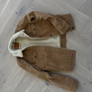 Levi’s denim jacka - Säljer en brun jeansjacka från Levi's med vitt teddyfoder på insidan och kragen. Jackan har klassiska jeansjacke-detaljer som bröstfickor med knappar och synliga sömmar. Perfekt för kyligare dagar. Några fläckar finns på ärmen samt ryggen, vet ej om de kan tvättad bort.