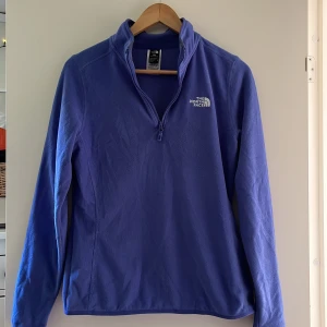 Blå fleecejacka från The North Face - Säljer en blå fleecejacka från The North Face med halv dragkedja och hög krage. Jackan har lång ärm och logga på bröstet samt ryggen. Perfekt för kyliga dagar och enkel att dra på över en t-shirt. Använts en gång!