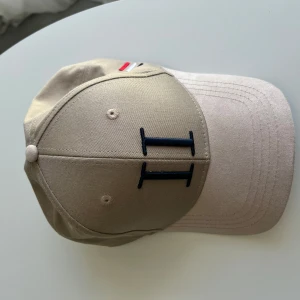 Beige keps från Les Deux - Snygg beige keps från Les Deux med justerbar baksida. Framsidan har en broderad svart 'II' och sidan har små broderade ränder i rött, vitt och blått. 
