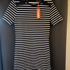 Randig klänning med spetsdetalj från Superdry - Mörkblå och vit randig klänning från Superdry med korta ärmar och spetsdetalj vid axlarna. Klänningen har en rak passform och är tillverkad i mjukt material, perfekt för en avslappnad stil.