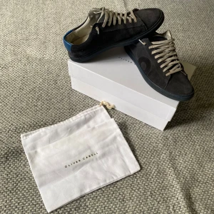 Svarta sneakers i mocka från Oliver Cabell - Säljer ett par svarta sneakers från Oliver Cabell i mocka med vita snören och blå detalj på hälen. Skorna har rund tå och platt sula, perfekt för en clean och stilren look. Levereras med originalpåse och låda. Storlek 41 men stora i storleken så mer som 42.