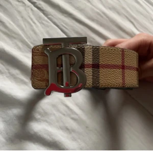 Burrberry bälte - Säljer ett beige bälte från Burberry med klassiskt rutigt mönster i rött och brunt. Bältet har ett stort silverfärgat spänne med Burberrys ikoniska 'TB'-logga. Perfekt accessoar för att lyfta din outfit. Går att byta mellan svart och biege sida jätte smidigt för att passa vilken outfit som helst 