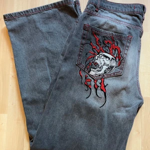 Dödskallejeans – Grå med röda detaljer - Unika grå jeansbyxor med röd kontrastsöm och stort broderat dödskallemotiv med eldflammor på bakfickan. Byxorna har dragkedja och raka, vida ben för en cool och avslappnad look.