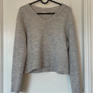 Ljusgrå stickad tröja från Gina Tricot - Mysig ljusgrå stickad tröja från Gina Tricot med v-ringad hals och ribbade detaljer vid ärmslut och nederkant. Tröjan har en något croppad passform och långa ärmar, perfekt för kyligare dagar.